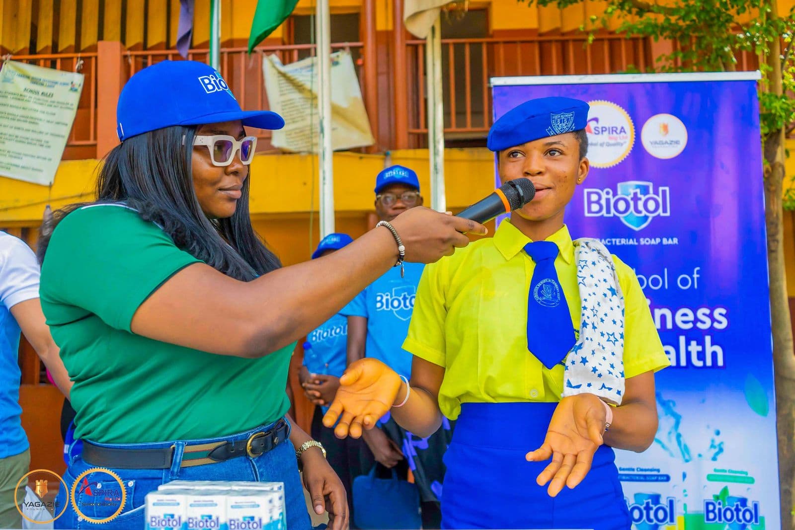 A facilitator in green demonstrates hand-washing steps to a student beside a hygiene banner
